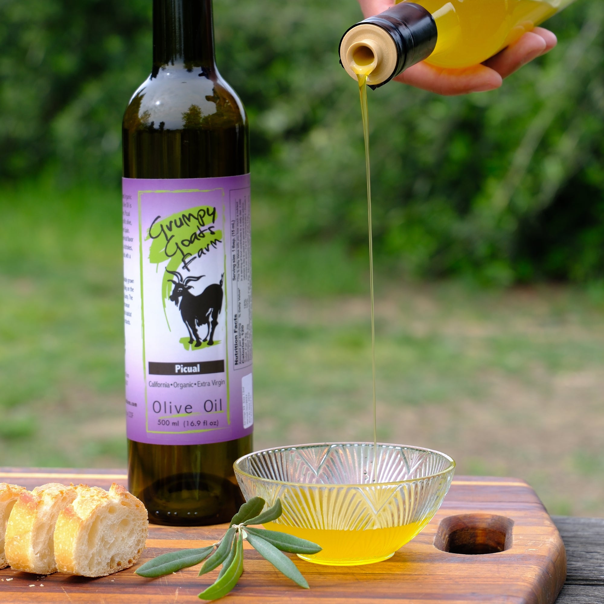 Grumpy Goats Organic Picual olive oil poured into a glass bowl beside bread and a 500 ml bottle.