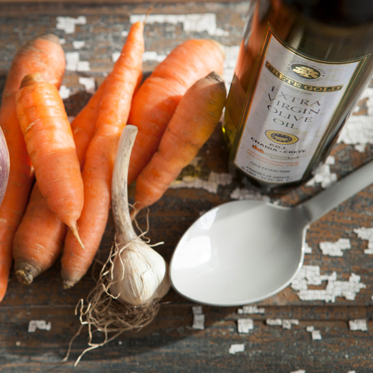 Crete Gold PDO Kolymvari olive oil beside fresh carrots, garlic and a serving spoon.