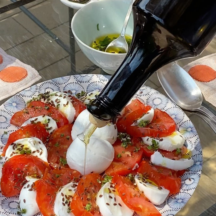 Marina Colonna extra virgin olive oil being poured over sliced tomatoes, mozzarella, herbs, and seeds.