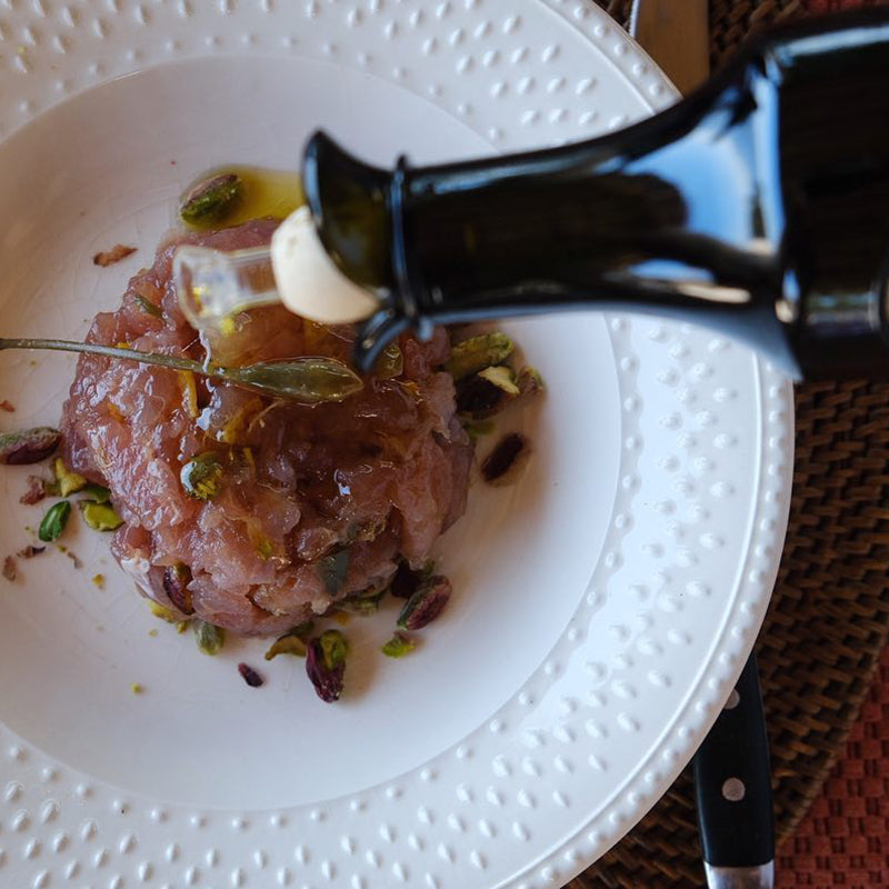 Colonna olive oil being poured over a plated raw fish dish with pistachios and capers.