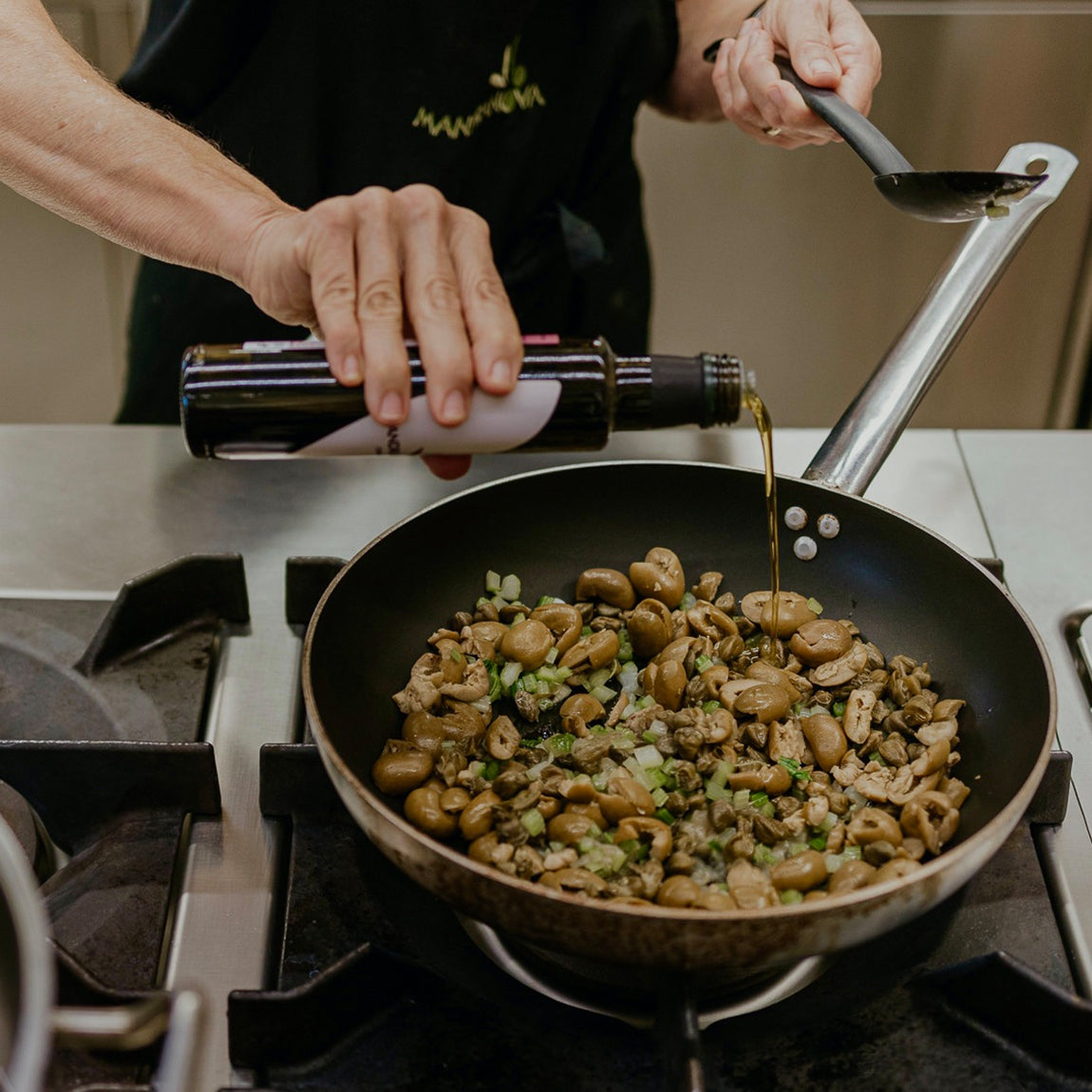 Pouring Mandranova Cerasuola olive oil into a frying pan filled with mushrooms, meat, and chopped vegetables.