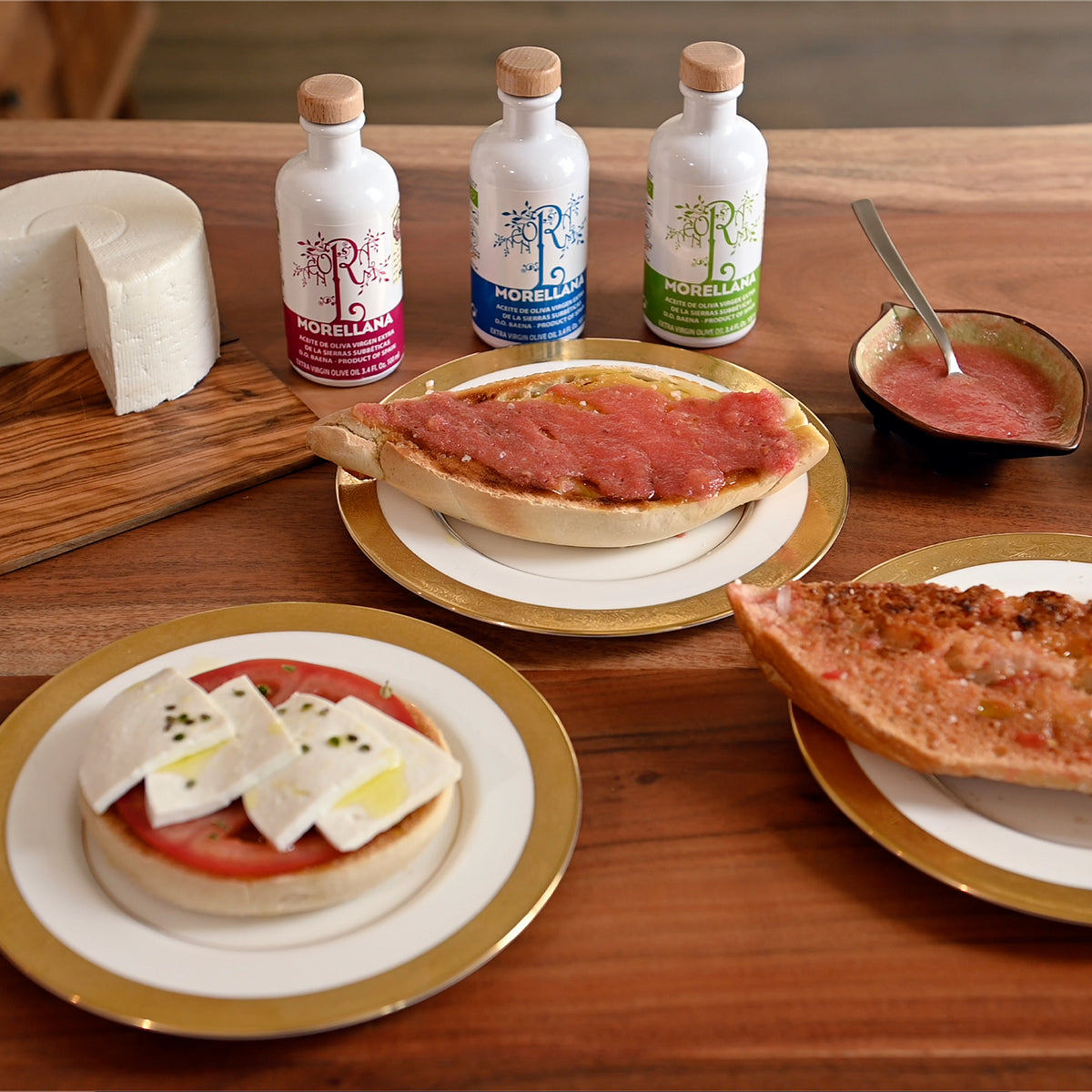 Three 100 ml Morellana organic olive oils beside cheese, tomato-topped bread, and plated appetizers.