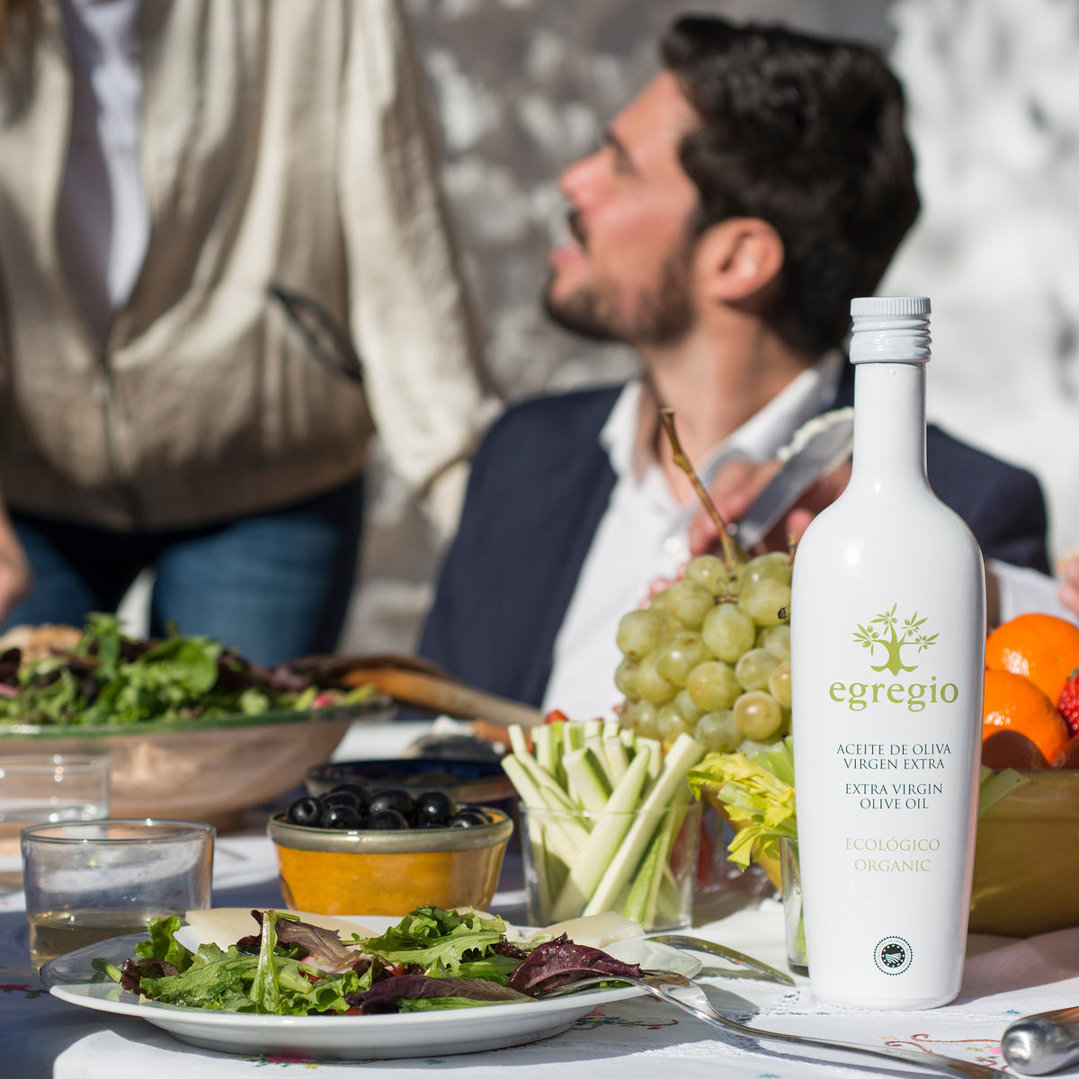 Oleoestepa Egregio Organic extra virgin olive oil on outdoor dining table with fresh salad, grapes and olives.