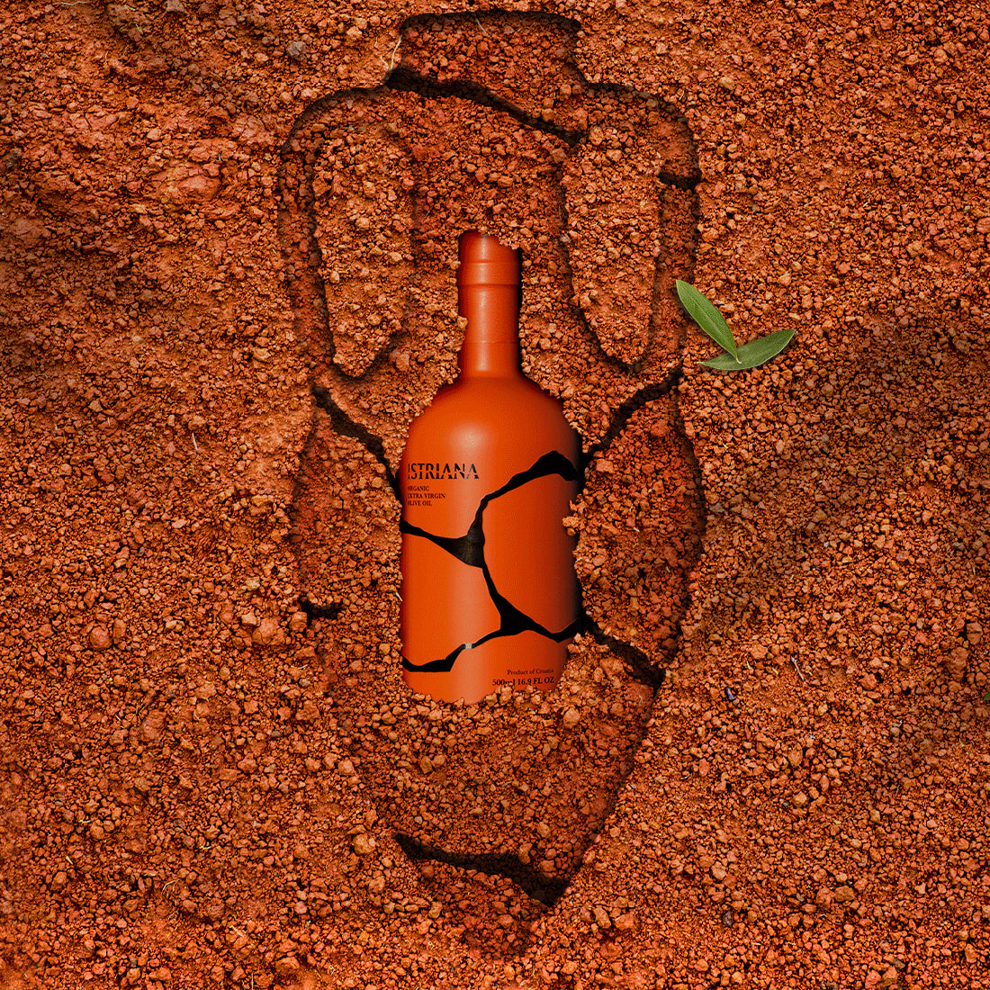 Istriana Organic 500 ml bottle nestled in reddish Istrian soil beside two green olive leaves.