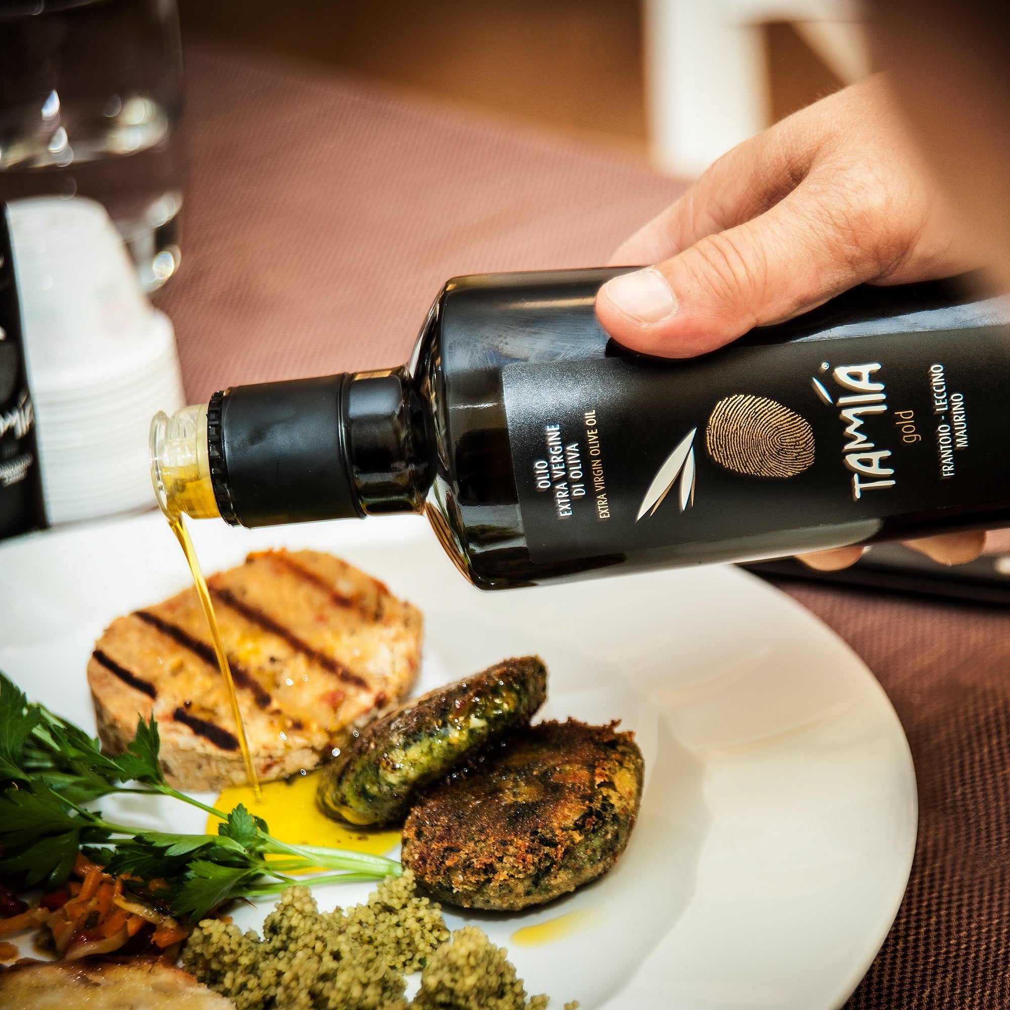 Tamia Gold 500 ml olive oil being poured over a plate of grilled bread, vegetables, and patties.