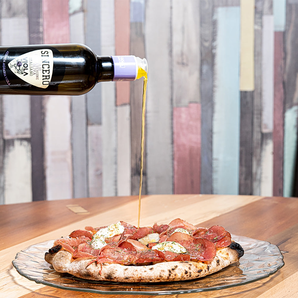 Viola Il Sincero extra virgin olive oil being poured over a prosciutto-topped pizza.