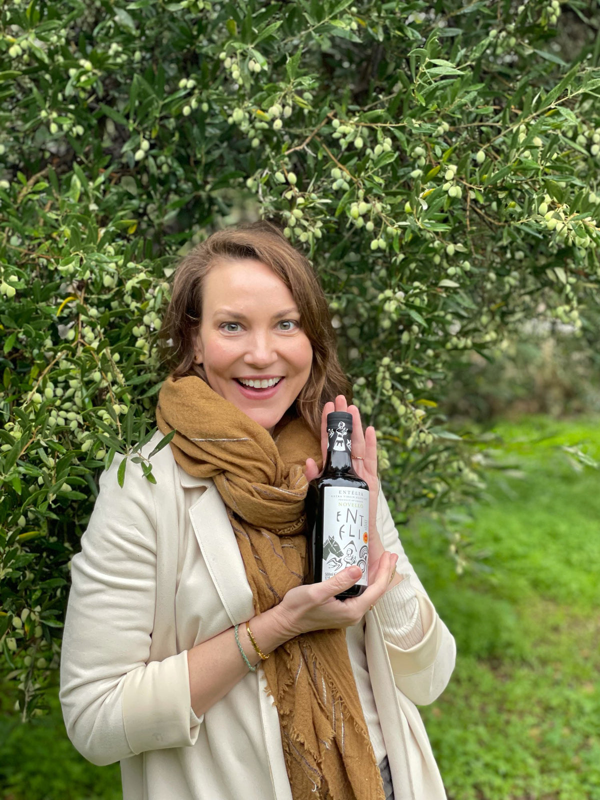 Olive Oil Lovers’ President and CEO, and olive oil expert, Joanne Lacina, holding a 500ml bottle of Entelia Novello Greek extra virgin olive oil near olive trees.